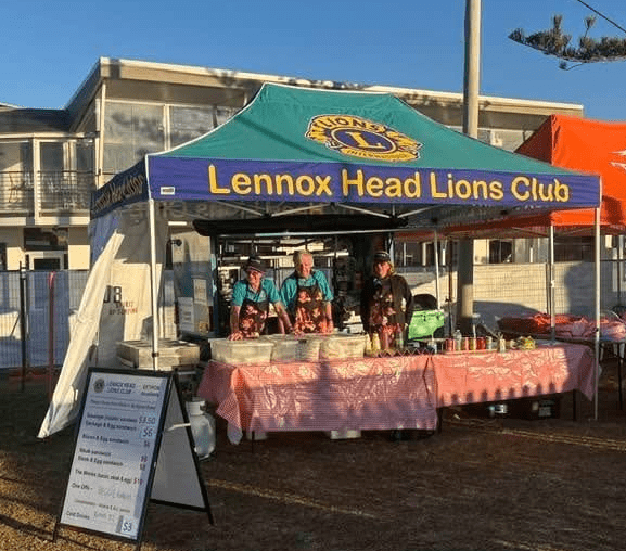 A group of Lions members in aprons over blue shirts manning a club BBQ stall under a branded awning.